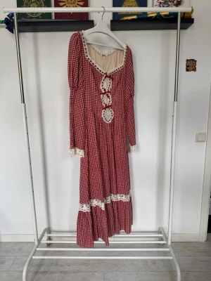 Röd långklänning från Lacemade  - En röd och vit rutig långklänning med spetsdetaljer vid halsringning, ärmslut och kjol. Klänningen kan knytas i ryggen för att dra in och framhäva midjan! Här är måtten från hemsidan! Bröst: 115cm, Midja: 95cm, Höft: 120cm