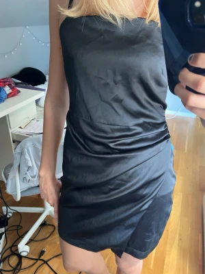Svart satin klänning med smala band - Snygg svart kort klänning i satin med smala axelband och rak urringning. Banden är påsydda, man kan klippa bort dem om man vill. Dragkedja där bak. Tyvärr är lappen borta då jag inte vet storlek eller märke, skulle säga att den är S