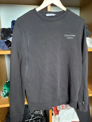 Svart sweatshirt från Calvin Klein Jeans - Snygg svart sweatshirt från Calvin Klein Jeans med diskret logga på bröstet. Tröjan har rund halsringning, ribbade muddar vid ärmslut och nederkant samt en klassisk passform. Perfekt för en stilren och avslappnad look. Storlek 16 år alltså xs till S