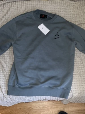 Grå crewneck sweatshirt från Jordan - Snygg grå crewneck sweatshirt från Jordan med svart Jumpman-logga och Nike Swoosh broderat på bröstet. Tröjan har ribbade muddar vid ärmslut och nederkant. Tillverkad i mjukt bomullsmaterial för skön passform. Perfekt för en avslappnad streetwear-look. 
