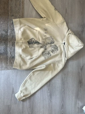 Beige zip hoodie med kors och tryck - Säljer en beige hoodie med dragkedja från Chrome Hearts. På framsidan finns ett stort svart kors-tryck med textdetaljer och små glittriga stenar. Hoodien har huva och två fickor framtill. Perfekt för dig som gillar streetwear och unika detaljer.