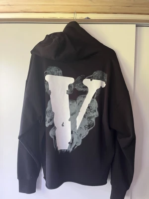 Svart Vlone hoodie med tryck - Svart hoodie från Vlone med stor vit V-logga och rökdetaljer på ryggen samt texten 'LONE' och en vinge på bröstet. Hoodien har huva med dragsko och ribbade muddar. Perfekt för dig som gillar streetwear och vill sticka ut.