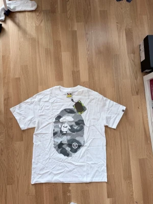 Vit BAPE t-shirt med camo-print - Säljer en vit t-shirt från BAPE med det ikoniska apansiktet i grått camo-mönster på bröstet. Klassisk passform och rund hals. Perfekt för dig som gillar streetwear och vill sticka ut med en unik design.