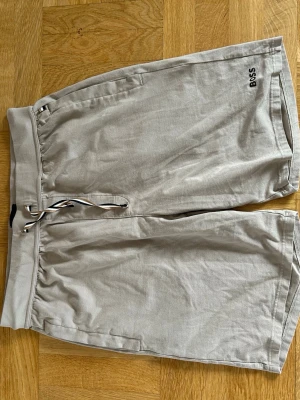 Beiga shorts från BOSS med snörning - Snygga beiga shorts från BOSS med bekväm midja och snörning framtill. Shortsen har två sidofickor och en diskret logga på vänster ben. Tillverkade i mjukt bomullsmaterial, perfekta för varma dagar.