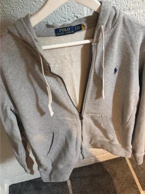 Grå zip hoodie från Polo Ralph Lauren - Snygg grå hoodie med dragkedja från Polo Ralph Lauren. Klassisk design med huva och vita snören. Skriv om ni har några frågor!!