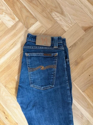 Blå jeans från Nudie Jeans - Säljer ett par klassiska blå  jeans från Nudie Jeans med snygga kontrastsömmar och femficksmodell. Jeansen har en läderpatch bak i midjan och är tillverkade i mjuk denim med stretch för extra komfort.
