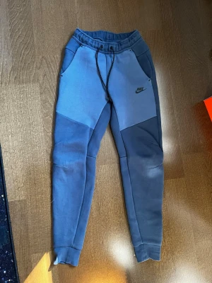 Nike Tech Fleece - Nike Tech i två nyanser av blått med elastisk midja och svartvit dragsko. Byxorna har sidofickor och Nike-logga tryckt på vänster lår. Tillverkade i mjukt och bekvämt material, perfekta för träning eller chill.