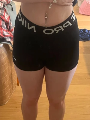 Svarta Nike Pro shorts - Säljer ett par svarta Nike Pro shorts med bred elastisk midja och vit logotyp. De är tighta och har en sportig look, perfekta för träning eller gym. Materialet är syntet och ger en bekväm passform som följer kroppen. Använda mycket och ända slitage som syns är märket på benet, annars är de som nya. 