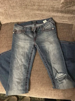 Blå jeans från True Religion - Säljer ett par blå jeans från True Religion med slitningar och hål på ena knät. Jeansen har klassiska röda sömmar, fem fickor och tydlig logga på bakfickan. Perfekta för en avslappnad och trendig look. Jättefina och super sköna. Priset går att diskutera 
