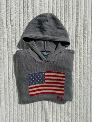 Grå hoodie från Polo Ralph Lauren - Snygg grå hoodie från Polo Ralph Lauren med stickad amerikansk flagga på bröstet och RL-broderi. Tillverkad i mjukt stickat material och har en klassisk huva. Perfekt för en avslappnad och cool stil.