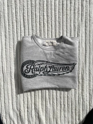Grå sweatshirt från Ralph Lauren - Snygg grå sweatshirt från Ralph Lauren med svart tryck på bröstet. Tröjan har rund halsringning, långa ärmar och ribbade muddar vid ärmslut och nederkant. Perfekt för en avslappnad och stilren look.