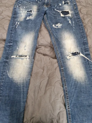 Blå slitna jeans med hål - Säljer ett par blå jeans med slitningar och stora hål på benen. Jeansen har en klassisk femficksmodell och är tillverkade i denim. De har en rak passform och tydliga slitdetaljer för en trendig look.