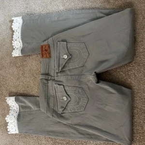 Gråa True Religion jeans med spets - Snygga gråa jeans från True Religion med raka ben och unika vita spetsdetaljer längst ner vid bensluten. Jeansen har två bakfickor med lock och knappar samt klassisk logopatch i läder bak. Perfekt för dig som vill sticka ut med din stil. Jeansen är använda endast en gång, men jag köpte de på Depart.A.Mental. På benet är de lite konstigt sydda, men det är ingen man tänker på. 💕