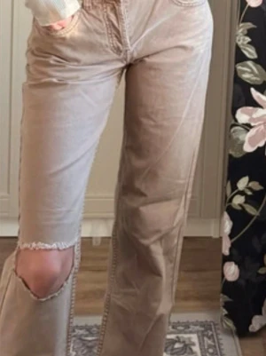 Beige raka jeans med hål på knät - Säljer ett par beige jeans med raka ben och ett stort hål på ena knät. Jeansen har hög midja, klassiska fickor både fram och bak samt råa kanter vid hålet. Perfekta för en avslappnad och trendig look.
