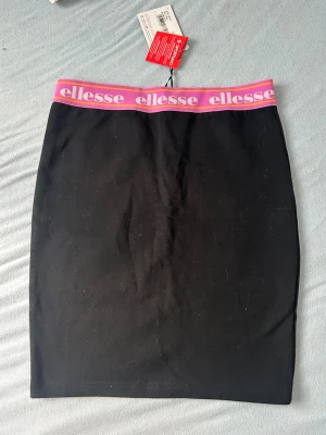 Svart kjol från Ellesse med logoband - Säljer en svart, tight kjol från Ellesse med ett brett elastiskt midjeband i rosa och lila med Ellesse-loggan runtom. Kjolen har en enkel, rak form och är perfekt för en sportig och trendig look.