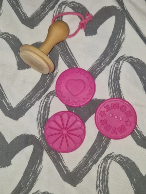 Kakstämpelset - Säljer ett stämpelset för kakor med ett handtag i trä och tre utbytbara stämplar i rosa silikon. Motiven är ett hjärta, en blomma och texten 'HOME MADE' med stjärnor. Perfekt för att göra personliga kakor eller bakverk.