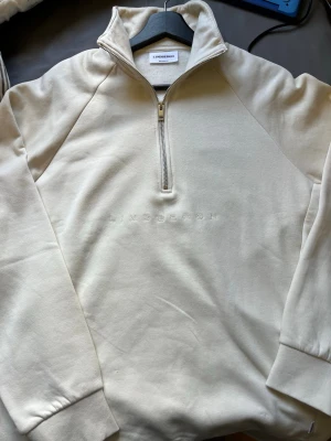 Beige halfzip sweatshirt Lindbergh - Stilren beige sweatshirt från Lindbergh med halv dragkedja och hög krage. Tillverkad i mjukt bomullsmaterial och har diskret broderad logga på bröstet. Långa ärmar med ribbade muddar ger en clean och modern look.