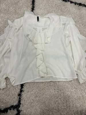 Vit blus med långa ärmar - Stilren vit blus med volangdetaljer längs kragen och framtill. Blusen har långa ärmar och detaljer. Tillverkad i ett tunt, mjukt tyg som ger ett elegant fall. Perfekt för sommaren. Skriv om du har frågor! 