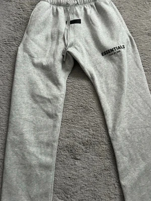 Grå mjukisbyxor Essentials Fear of God - Snygga grå mjukisbyxor från Fear of God Essentials med svart logga tryckt på låret. Byxorna har en klassisk rak passform och är gjorda i mjukt och bekvämt material, perfekt för en avslappnad stil.