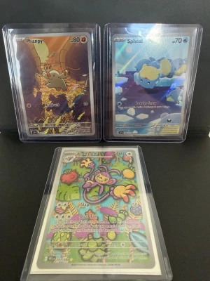 Pokémonkort Phanpy, Spheal & Ambipom - Tre Pokémonkort i plastfickor: Phanpy med orange bakgrund, Spheal med blått ismotiv och Ambipom med färgglad, tropisk design. Alla kort har detaljerade illustrationer och tydliga Pokémon-namn. Perfekt för samlare eller Pokémonfans.