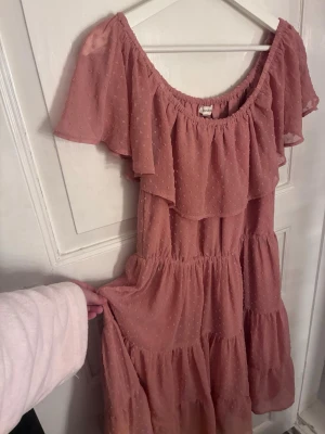 Rosa volangklänning från Newbie - Söt rosa klänning från Newbie med volanger och prickigt mönster. Klänningen har korta ärmar, en luftig passform och omlottvolang upptill. Perfekt för sommarens alla tillfällen och har en romantisk känsla.