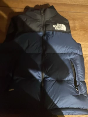Blå och svart dunväst The North Face - Snygg blå och svart dunväst från The North Face med dragkedja framtill och två sidofickor. Västen har en klassisk puffer-stil och är perfekt för lager-på-lager. Tydlig logga på bröstet och hög krage för extra värme.