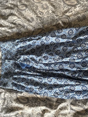 Blå mönstrad långkjol med knyt - Säljer en blå långkjol med vitt och mörkblått blommigt mönster. Kjolen har smock i midjan och dekorativa knytband med tofsar framtill. Luftigt material som passar perfekt till varma dagar.
