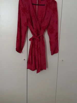 Röd omlottklänning med knytband och shorts - Snygg röd omlottklänning med knytband i midjan och långa ärmar. Klänningen har subtilt blommönster och djup v-ringning, perfekt för dig som vill sticka ut. Lätt och skönt material som ger en feminin siluett.
