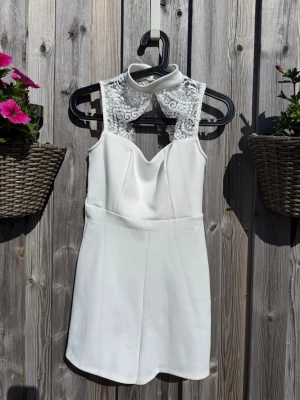 Vit spetsjumpsuit med hög krage (XS) - Supersnygg vit jumpsuit med hög krage och detaljer i spets upptill. Ärmlös modell med hjärtformad urringning och öppen rygg med spetsmönster. Perfekt för dig som vill sticka ut lite extra. Materialet känns lätt och luftigt.