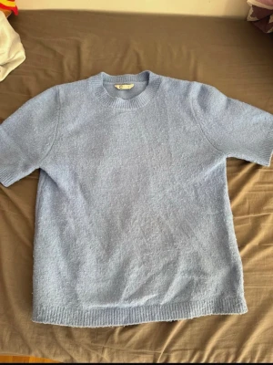 Ljusblå stickad t-shirt - Ljusblå stickad t-shirt med korta ärmar och rund hals. Tröjan har ribbade muddar vid hals, ärmslut och nederkant. Perfekt för dig som gillar en enkel men stilren look. Materialet ser mjukt och bekvämt ut. Lite nopprig!