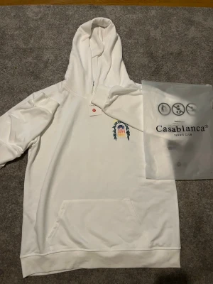 Vit hoodie Avenida Casablanca - Vit hoodie från Avenida med färgglad tryck på både fram- och baksidan. Motivet visar en fönsterram med gröna blad och en byggnad under en natt- och solhimmel. Hoodien har huva och en stor magficka. Perfekt för dig som gillar unika prints.