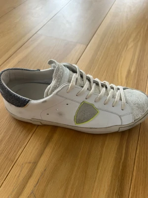 Vita Philippe Model sneakers med patch - Snygga vita sneakers från Philippe Model med grå mockadetaljer på tå och plös, samt en grå patch med gul kant på sidan. Hälpartiet har en mörkgrå textilpanel med struktur. Klassisk låg modell med vita skosnören och gummisula.