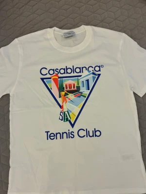Vit Casablanca Tennis Club t-shirt - Snygg vit t-shirt från Casablanca med färgglatt tryck och texten 'Tennis Club' på framsidan. Klassisk passform och rund hals. Perfekt för dig som gillar sportig och trendig stil.