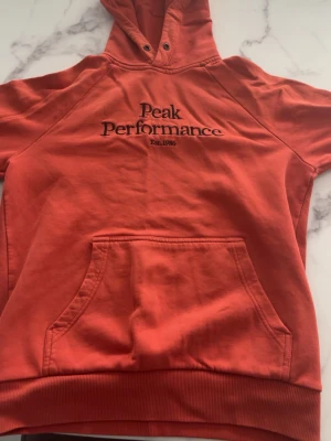 Röd/orange hoodie från Peak Performance - Snygg röd/orange hoodie från Peak Performance med broderad logga och text 'Est. 1986' på bröstet. Klassisk känguruficka framtill, ribbade muddar och huva med dragsko. Perfekt för en avslappnad och sportig stil.