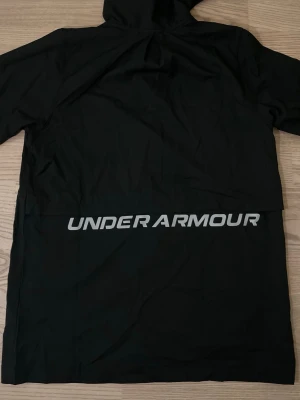 Svart vindjacka från Under Armour - Svart vindjacka med huva från Under Armour. Jackan har dragkedja framtill, vita detaljer vid axlarna och logga på bröstet. Stor Under Armour-tryck på ryggen. Lätt och sportig design, perfekt för blåsiga dagar.