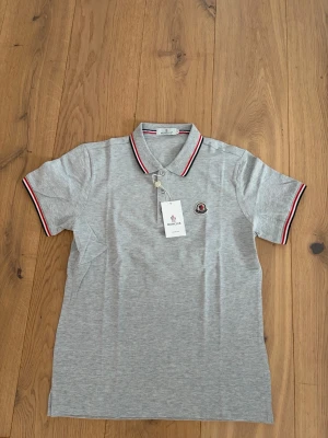 Grå Moncler piké t-shirt - Snygg grå piké t-shirt från Moncler med klassisk krage och knappar framtill. T-shirten har röda och vita detaljer på krage och ärmslut samt Moncler-logga på bröstet. Perfekt för en stilren och sportig look.