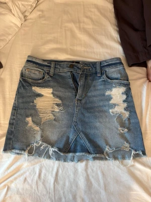 Blå jeanskjol från Hollister - Säljer en blå jeanskjol från Hollister med hög midja och slitna detaljer framtill. Kjolen har klassisk femficksdesign, bälteshällor och rå kant nedtill. Perfekt för dig som gillar en avslappnad och trendig stil.