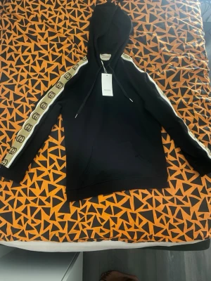 Svart hoodie Gucci Reflective  - Svart hoodie från Gucci med huva och dragsnören. Längs ärmarna går ett beige band med klassiska Gucci-loggor i brunt. Hoodien har en avslappnad passform och är tillverkad i mjukt material, perfekt för en trendig och lyxig streetwear-look.