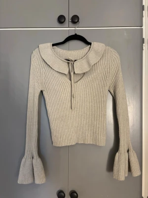 Beige ribbstickad tröja med krage - Trendig beige ribbstickad tröja med stor krage och knytband framtill. Långa ärmar med volangdetalj vid ärmslut ger en unik look. Perfekt för dig som gillar en söt och stilren stil. Materialet känns mjukt och följsamt.