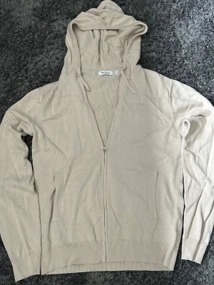 Beige hoodie med dragkedja från RVLT - Snygg beige hoodie från RVLT med dragkedja framtill och två fickor.Storlek 170-178. Tillverkad i ett mjukt, tunt material som känns skönt mot huden. Klassisk huva med dragsko och ribbade muddar vid ärmslut och nederkant. Perfekt för en avslappnad stil.