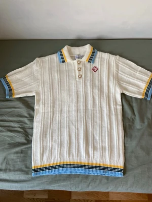 Casablanca  - Vit ribbad henley från Casablanca med korta ärmar och färgglada ränder i blått och gult på krage, ärmslut och nederkant. Skjortan har knappar framtill och en liten broderad logga på bröstet. Perfekt för en sportig och retro look.