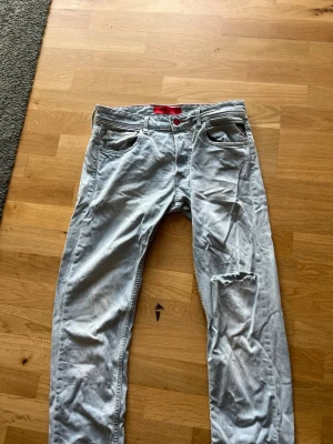 Ljusblå jeans från Hugo Replay - Säljer ett par ljusblå jeans från Hugo Replay med rakt ben och slitning på höger knä. Jeansen har klassisk femficksdesign, röda detaljer och är tillverkade i denim. Perfekta för en avslappnad och trendig look. Som ni ser så är det en mindre fläck vid gylfen som ja it riktigt kunnat få bort. Vid funderingar så skriv ba