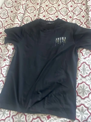 Svart AMIRI t-shirt med tryck - Snygg svart t-shirt från AMIRI med färgstarkt tryck på bröstet där märkesnamnet syns med droppande effekt i flera färger. Klassisk passform och rund halsringning, perfekt till jeans eller shorts. Tillverkad i mjuk bomull för skön känsla.