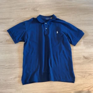 Marinblå pikétröja från Polo Ralph Lauren - Klassisk marinblå pikétröja från Polo Ralph Lauren med korta ärmar och vit broderad logga på bröstet. Tröjan har krage och två knappar framtill. Perfekt för en stilren och avslappnad look.