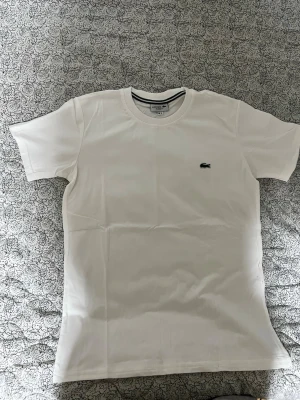 Vit t-shirt  - Klassisk vit t-shirt med rund halsringning och den ikoniska krokodilloggan broderad på bröstet. Tillverkad i mjuk bomull och har en normal passform med korta ärmar. Perfekt basplagg till garderoben.
