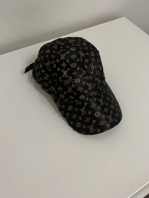 Svart keps med L och V mönster - Snygg svart keps  med det klassiska L och V monogrammet i beige över hela kepsen. Justerbar rem bak för perfekt passform. Tillverkad i ett glansigt material som ger en exklusiv känsla och sticker ut i mängden.