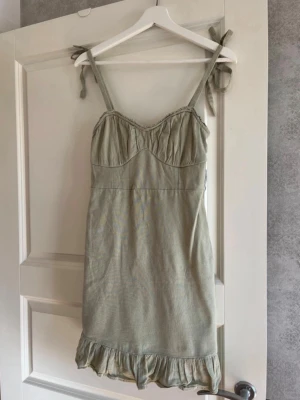 Ljusgrön klänning Hollister S - Supersöt ljusgrön klänning från Hollister med knytbara axelband och volangkant nertill. Klänningen har figurnära passform och smock i ryggen för extra komfort. Perfekt för varma dagar och enkel att styla med sneakers eller sandaler.