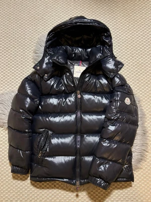 Svart Moncler Maya - Snygg svart dunjacka från Moncler med glansigt material och avtagbar huva. Jackan har en klassisk puffer-design, dragkedja framtill och Moncler-logga på ärmen. Perfekt för kalla dagar och riktigt stilren att bära. 