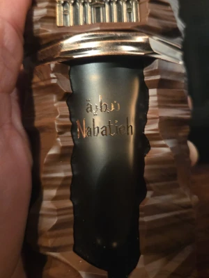 Nabatieh 100ml full - Unik parfymflaska från Nabatieh med en cool, skulpterad design i brunt och svart. Flaskan har gulddetaljer och en text i guld på framsidan. Perfekt för dig som gillar exklusiva och annorlunda dofter.