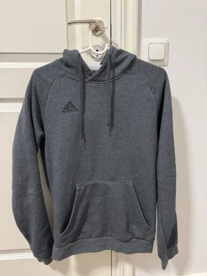 Grå hoodie från Adidas - Snygg grå hoodie från Adidas med klassisk logga på bröstet. Hoodien har huva med dragsnören och en stor magficka framtill. Tillverkad i mjukt material som känns skönt mot huden. Perfekt för en avslappnad och sportig stil.